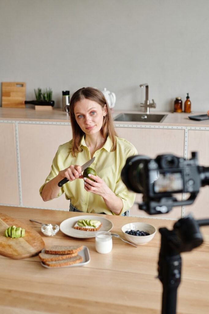 Caucasian woman demonstrating avocado preparation for a vlog in a modern kitchen setting.
